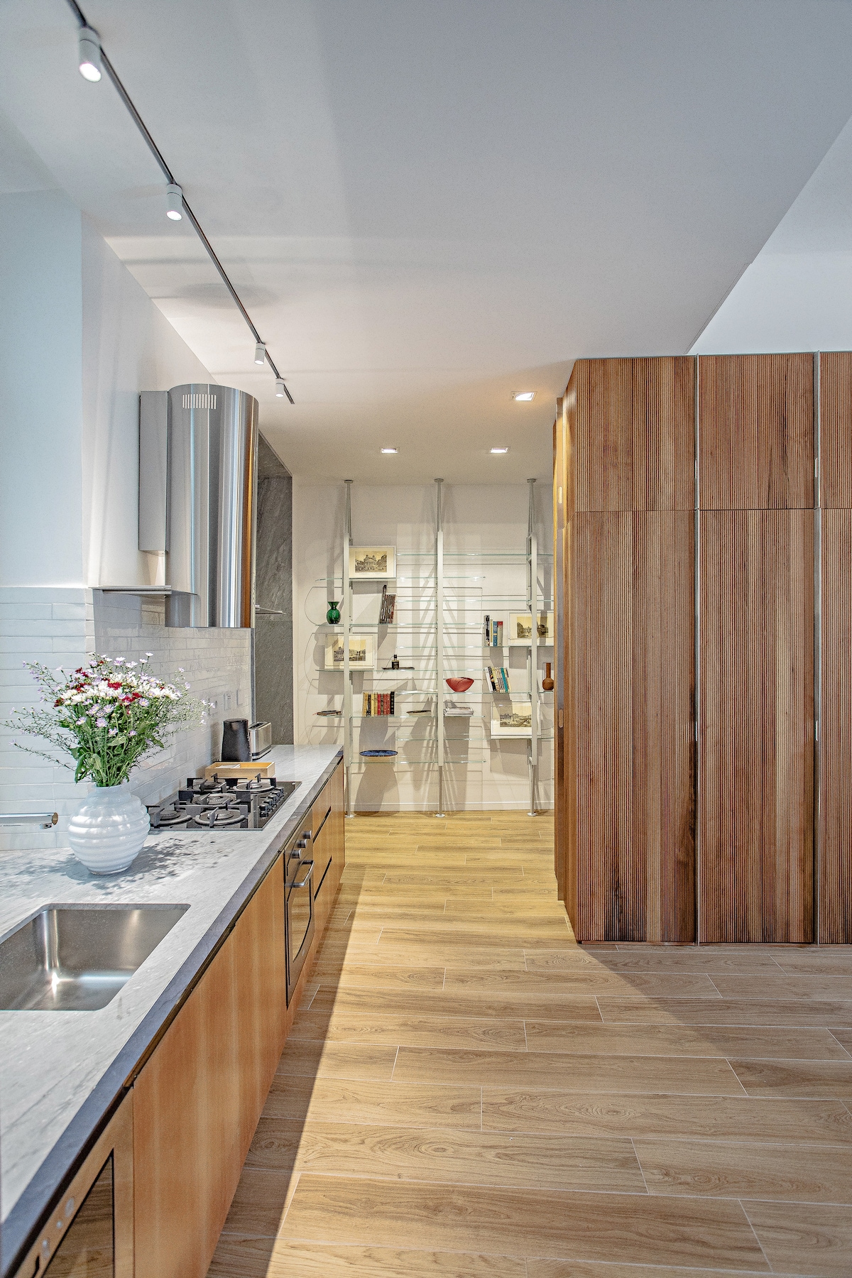 A modern kitchen features a sleek countertop and stainless steel appliances, with wooden cabinetry enhancing the design. Soft lighting illuminates the space, highlighting the light-colored walls and the organized shelving area in the background.