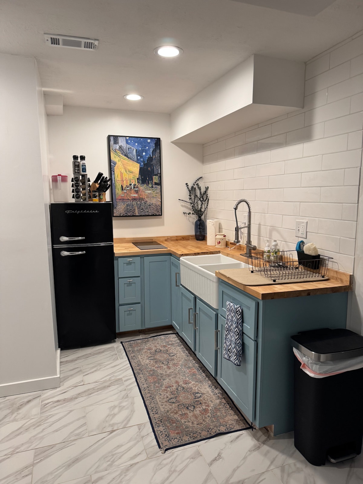 A compact kitchen area is designed with blue cabinetry and a wooden countertop. A sink is situated beside a black refrigerator. Decorative items, including a vase and a wall art piece, add character. A small rug lies on the tiled floor, complementing the overall design.