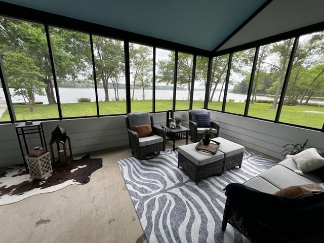 A screened-in porch features comfortable seating arrangements on a patterned rug, complemented by a small table at the center. Large windows offer unobstructed views of the surrounding greenery and tranquil lake, creating a serene spot for relaxation.