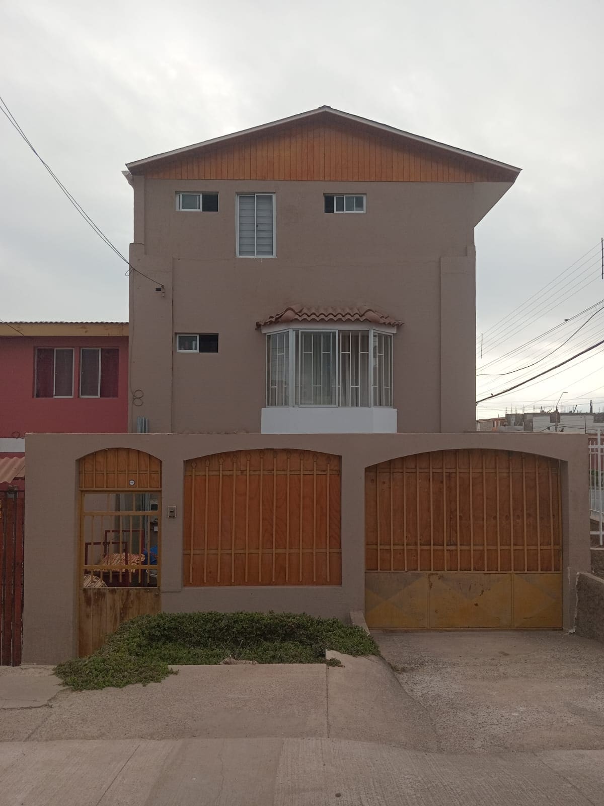 A three-story building with a mix of neutral and wooden siding is depicted. The entrance features two wooden gates, while a balcony is visible on the second floor. Windows are positioned thoughtfully, allowing natural light to enter. Surroundings include city infrastructure with power lines.