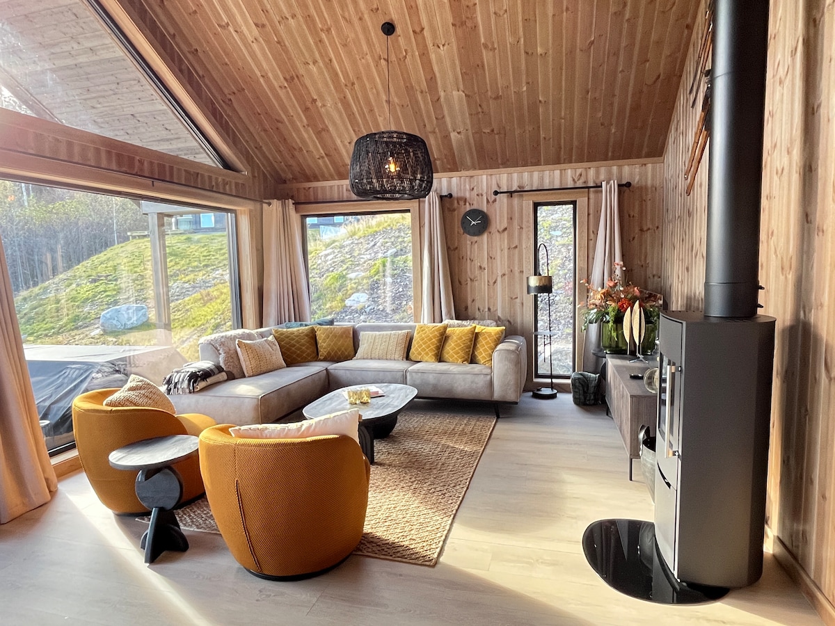 A modern living area features large glass doors that open to the outdoor landscape. A spacious corner sofa in light fabric is accompanied by two bright orange chairs. Natural light fills the room, highlighting a wood-burning stove and decorative plants on a side table.