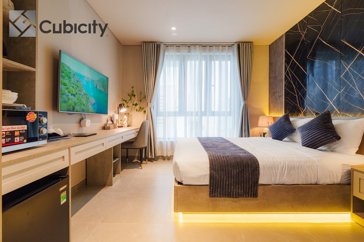 A modern bedroom is presented, featuring a spacious bed with a dark throw and pillows. A wide window allows natural light to enter, while a television is mounted on the wall. A desk, mini-fridge, and microwave are included in the cozy, well-organized space.