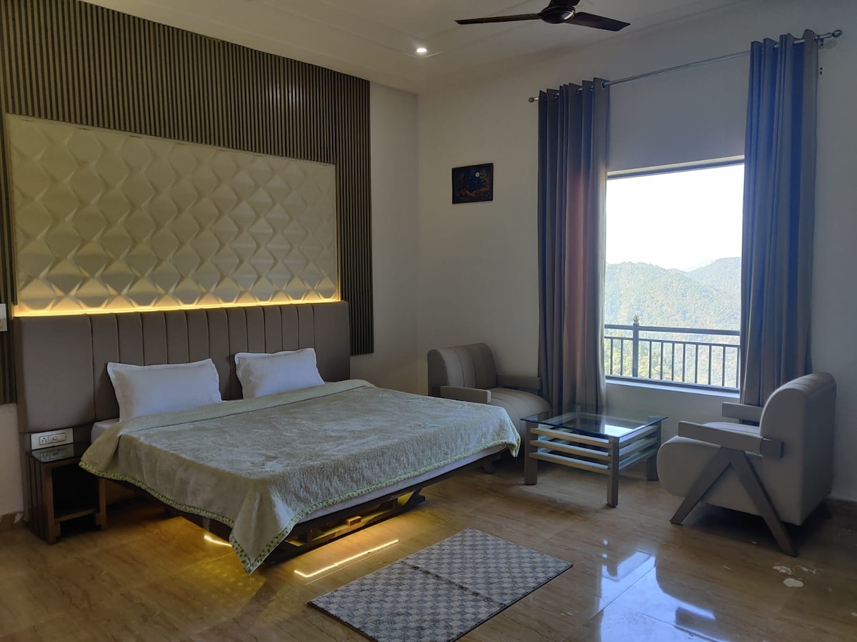 A spacious family suite features a large bed with neutral linens. A small seating area includes a modern chair and a glass-top table. Natural light enters through a large window with light curtains, providing a view of distant hills. The room's design incorporates soft lighting and textured wall panels.