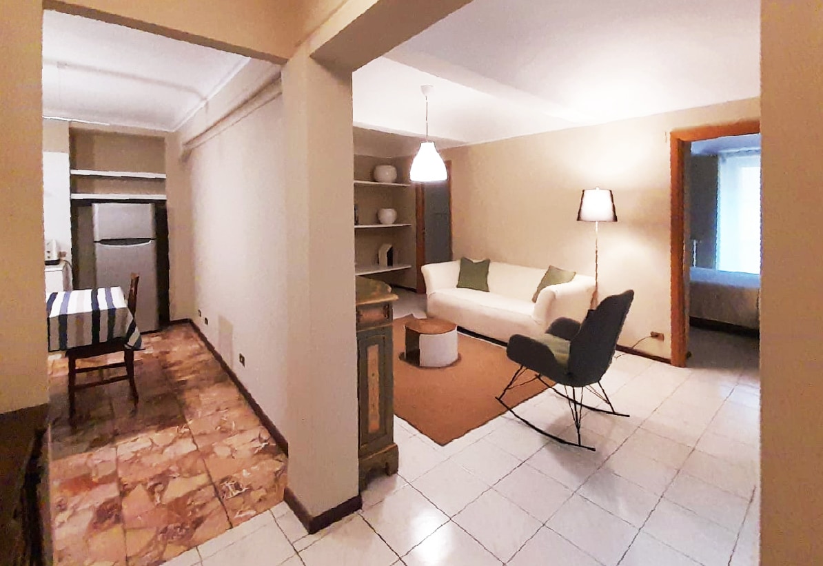 An open layout is presented with a living area featuring a light-colored sofa and accent chair, complemented by a neutral rug. A dining table with a striped tablecloth is visible in the background, along with a simple shelving unit and an inviting light fixture overhead.
