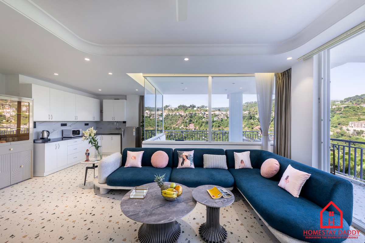 An open living area features a plush, curved sectional sofa adorned with decorative pillows. A round coffee table sits between the couch and a window, showcasing scenic views. Modern cabinetry and kitchen appliances are visible in the background, enhancing the space's contemporary feel.