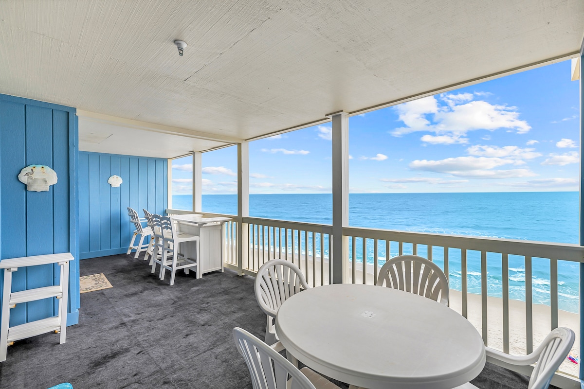 A spacious balcony features white outdoor furniture, including a round table surrounded by chairs. The ocean is visible beyond the railing, with waves gently lapping at the shore. Calm blue skies and soft clouds create a serene backdrop.