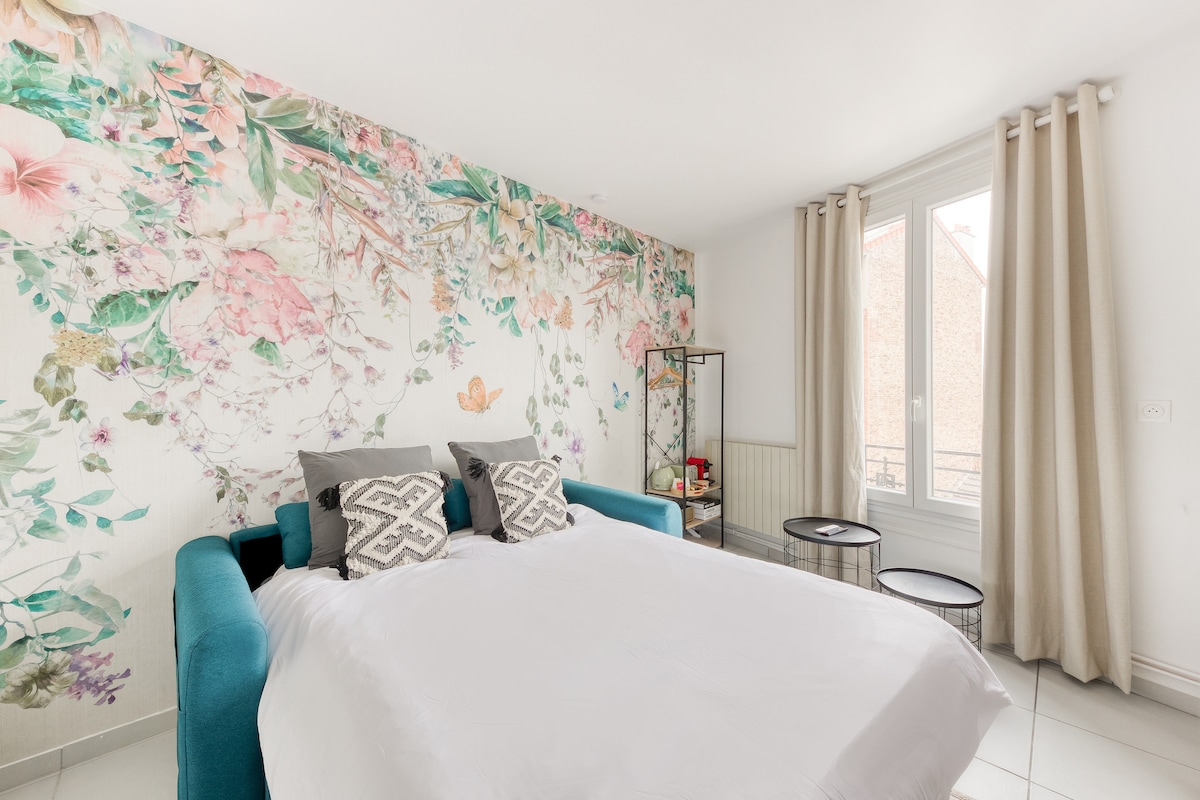 The sleeping area features a spacious bed with a white coverlet, accented by two patterned pillows. A floral mural adorns the wall behind the bed, while a stylish metal rack and two small tables add functionality. Natural light filters through the window, covered by light curtains.