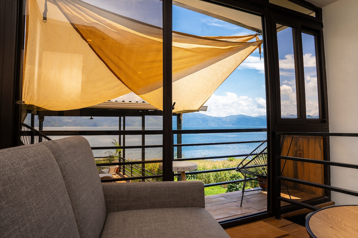 A comfortable seating area is visible through large glass doors, framed by a flowing yellow fabric. The serene view of the lake and surrounding mountains creates a peaceful backdrop, while the outdoor deck extends invitingly beyond the railing.