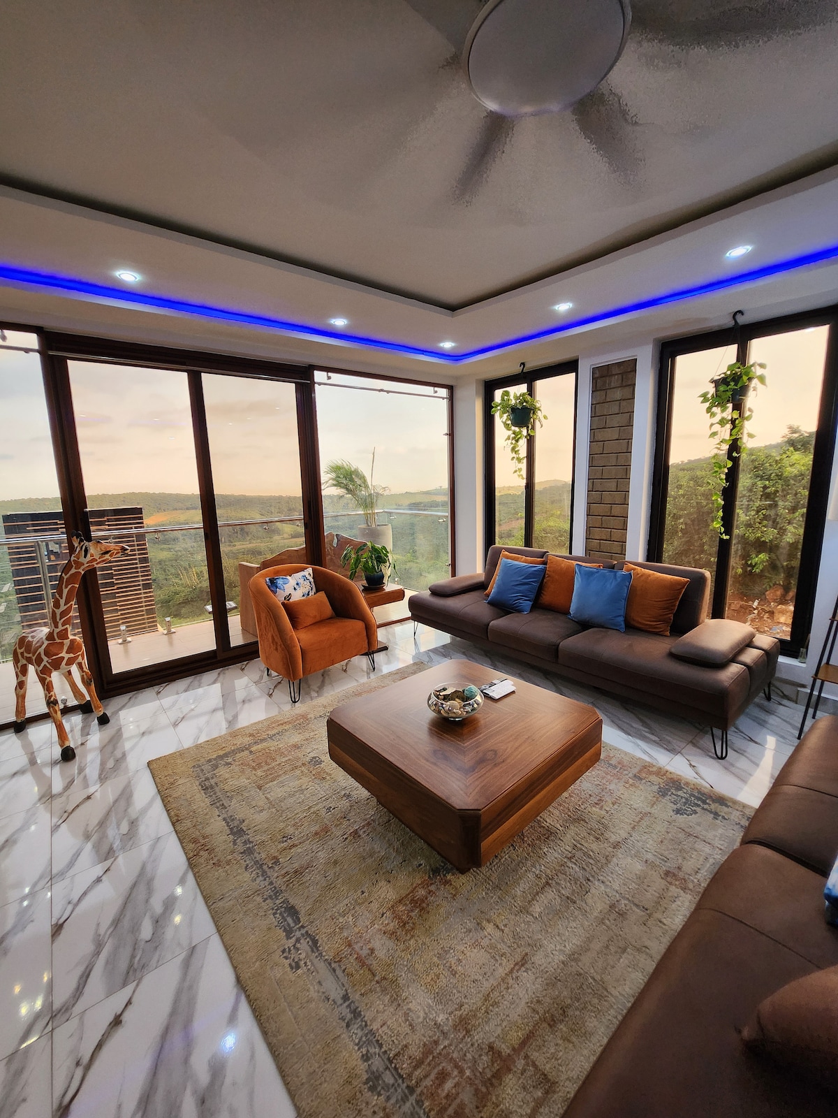 A modern living area is showcased with large windows that offer expansive views of the surrounding hills. A plush brown sofa is accompanied by an orange armchair. Decorative plants hang from the ceiling, and a wooden coffee table sits centrally on a patterned rug.
