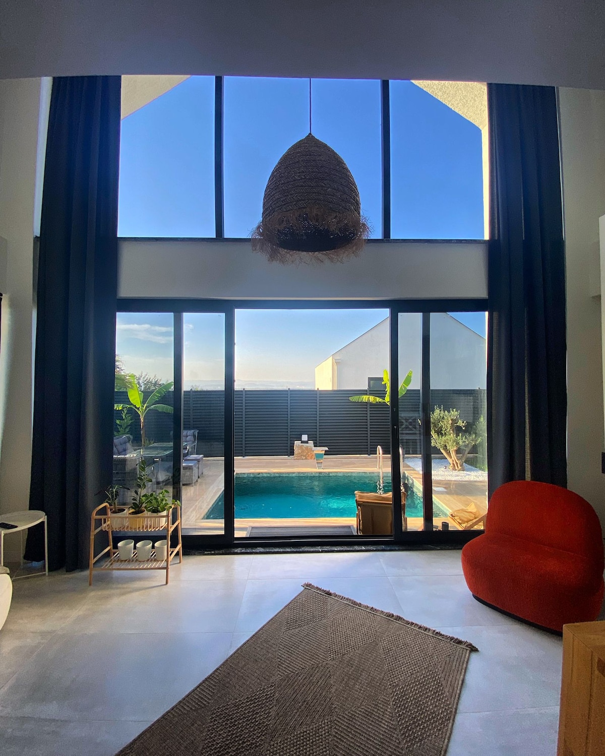 Large glass doors frame a view of the outdoor pool area, allowing natural light to fill the room. A modern pendant light hangs from the ceiling, complementing the plush seating area with a bold red accent chair. Indoor plants add greenery to the space.