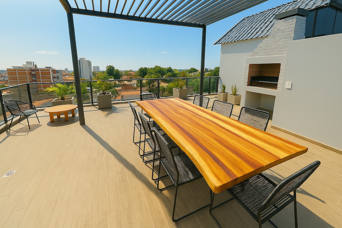 A spacious terrace features a long wooden dining table surrounded by black wire chairs. A barbecue area is visible to one side, and potted plants are arranged along the railing, providing a welcoming atmosphere with views of the surrounding area.