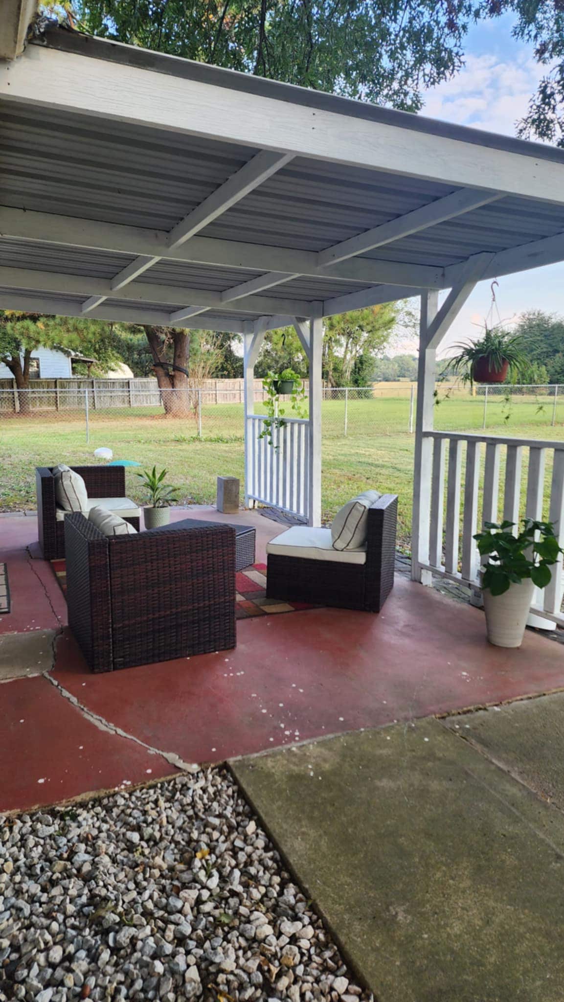 An outdoor seating area is present, featuring two woven armchairs and a matching coffee table on a shaded patio. Surrounding greenery creates a peaceful atmosphere, with gravel and potted plants adding texture to the space. A clear view of open pasture can be seen beyond the patio.