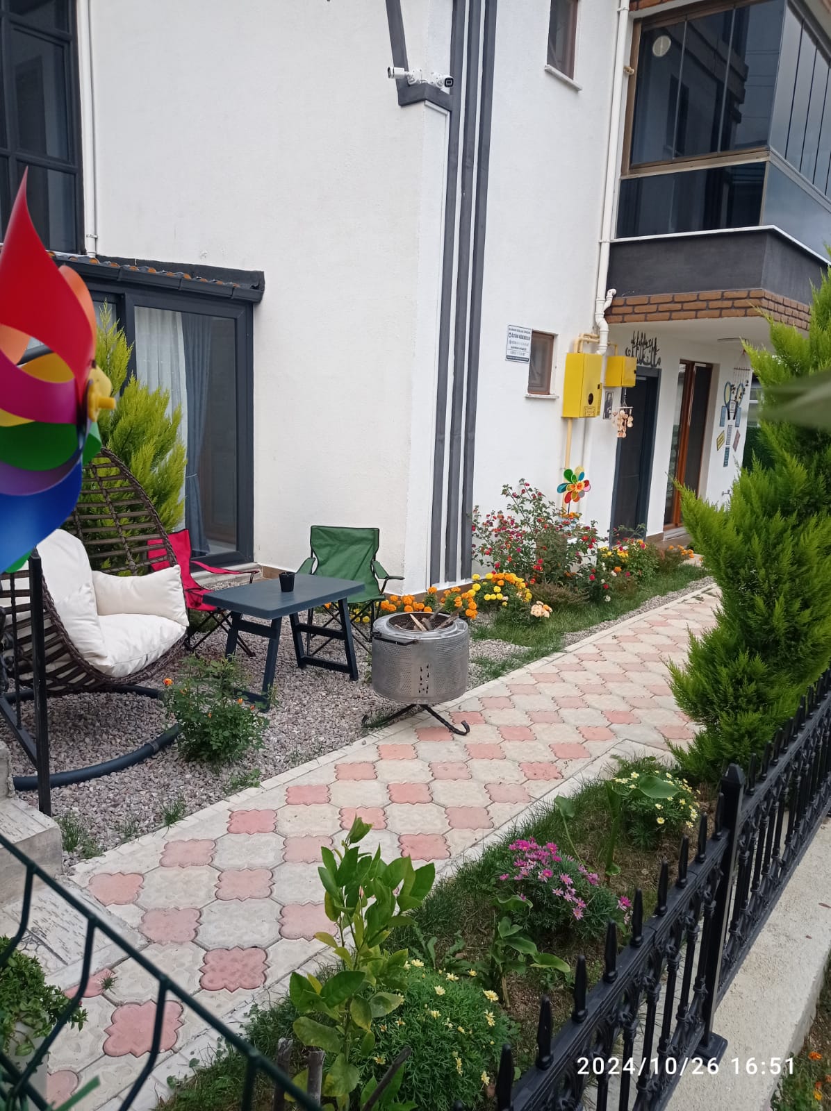 A charming garden area is displayed with a stone pathway lined by flowering plants and greenery. Seating is provided by a hanging chair and a small table. A colorful pinwheel adds a playful touch, while the exterior of the house offers a clean, modern aesthetic.