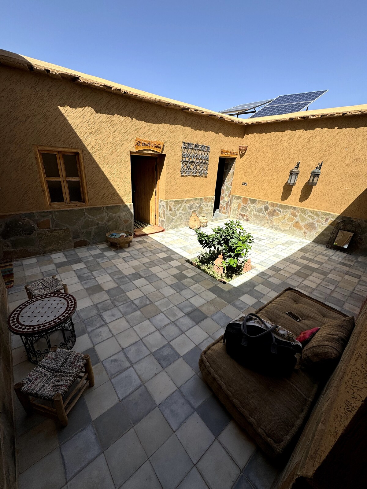 A sunlit courtyard features stone flooring, adorned with a small green plant and decorative elements. Rustic doors lead to adjoining rooms, and a comfortable seating area with woven accents is visible, inviting relaxation in this tranquil space.
