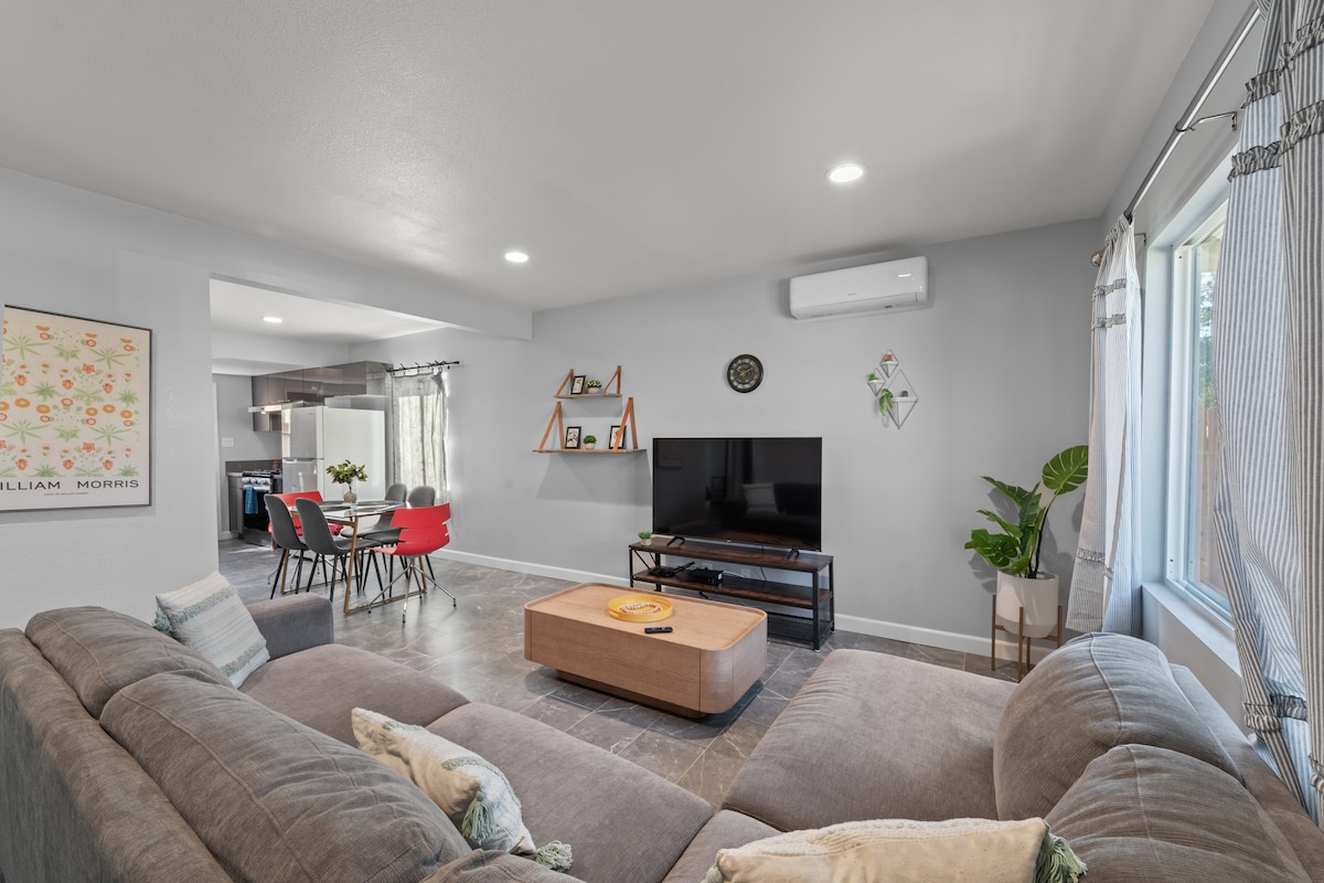 A comfortable living area features a spacious sectional sofa facing a flat-screen television. Natural light enters through a large window, illuminating the neutral grey walls. A wooden coffee table and decorative plants add a touch of warmth, while a dining area is visible in the background.
