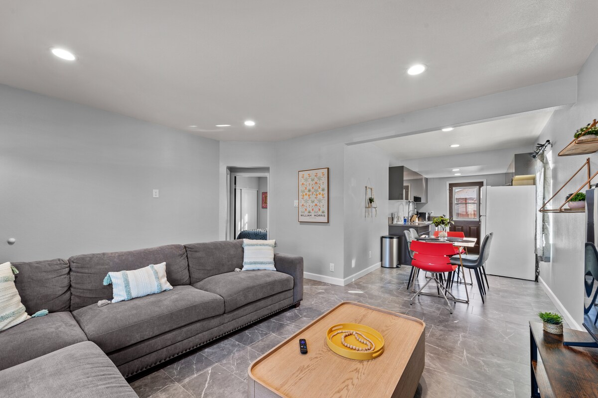 An open living area features a comfortable grey sectional sofa with pastel accent pillows. A wooden coffee table sits in front. The layout leads to a modern dining space with red chairs, visible through a doorway connecting to the kitchen.