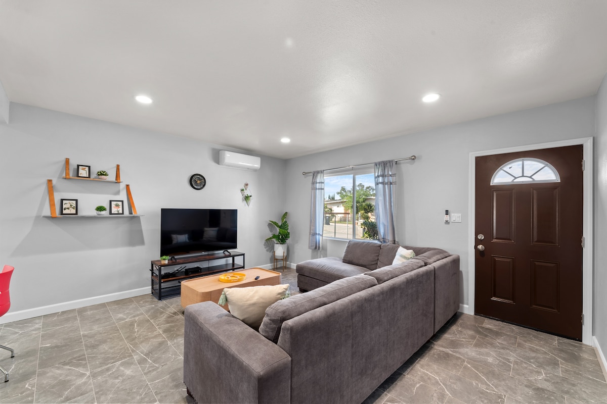 A spacious living area features a comfortable gray sectional sofa and large flat-screen television. Decorative shelves hold small plants and framed pictures. Natural light flows in through a window adorned with light curtains, and a welcoming front door adds to the inviting ambiance.