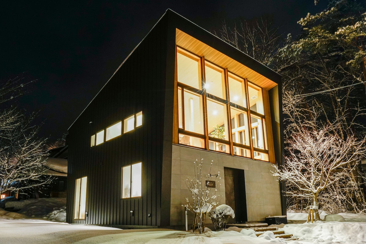 A modern home with a sleek black exterior is illuminated at night, showcasing large glass windows that provide a warm glow from within. Snow blankets the surrounding landscape, while trees with frosted branches frame the property, creating a serene winter scene.