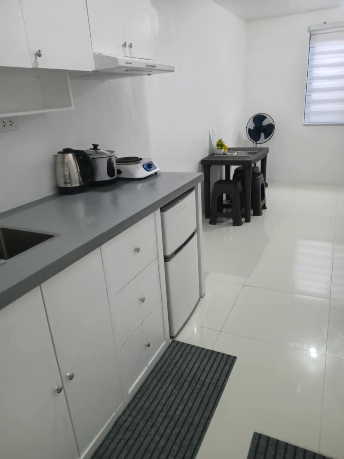 A well-equipped kitchen area is shown, featuring sleek cabinetry and a countertop with a sink. Appliances, including a kettle and a cooking device, are visible. A small dining table with stools is positioned nearby, and a fan provides additional comfort in the space.