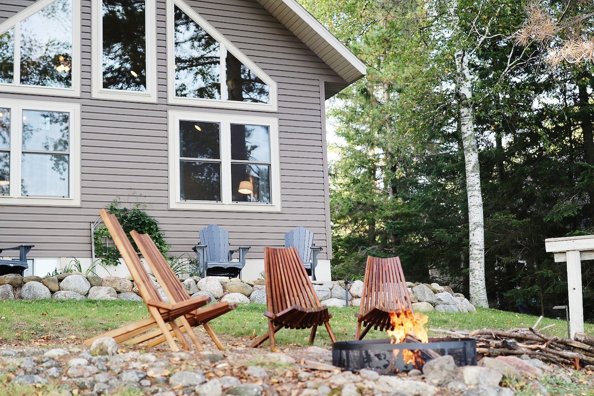 A fire pit is presented in the foreground, surrounded by three wooden lounge chairs. In the background, the exterior of a modern home is visible, featuring large windows that invite natural light. A pared stone landscaping complements the natural setting of trees.
