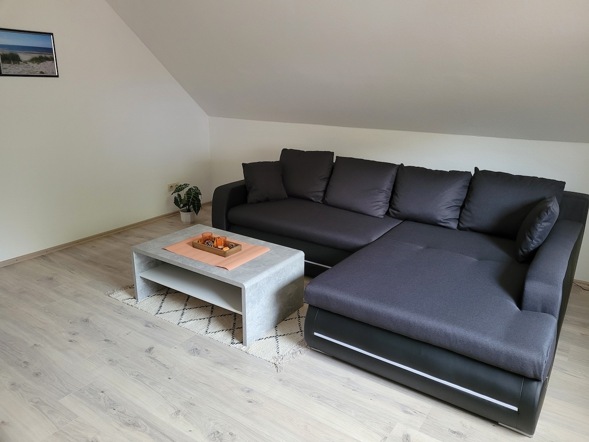A modern living area features a large sectional sofa in dark fabric, complemented by a minimalist coffee table made of concrete. A decorative tray sits atop the table, and a small plant adds a touch of greenery to the space. Natural light fills the room from a nearby window.