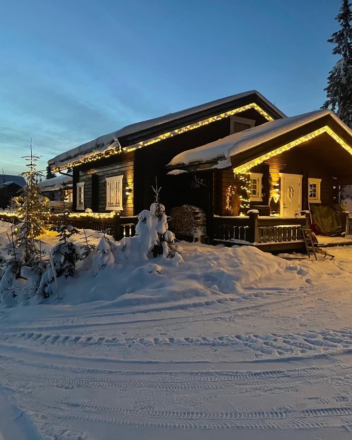 A cozy black wooden cabin is partially covered in snow, with warm lights outlining the roof. Surrounding trees are blanketed in white, creating a serene winter scene. A snow-covered pathway leads to the entrance, inviting guests to explore the charming atmosphere.