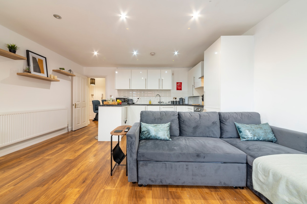 An open-plan living area presents a modern design with a comfortable sofa adorned with cushions. The bright kitchen features contemporary cabinetry and a sleek countertop. Hardwood flooring enhances the space, and recessed lighting creates a warm ambiance.