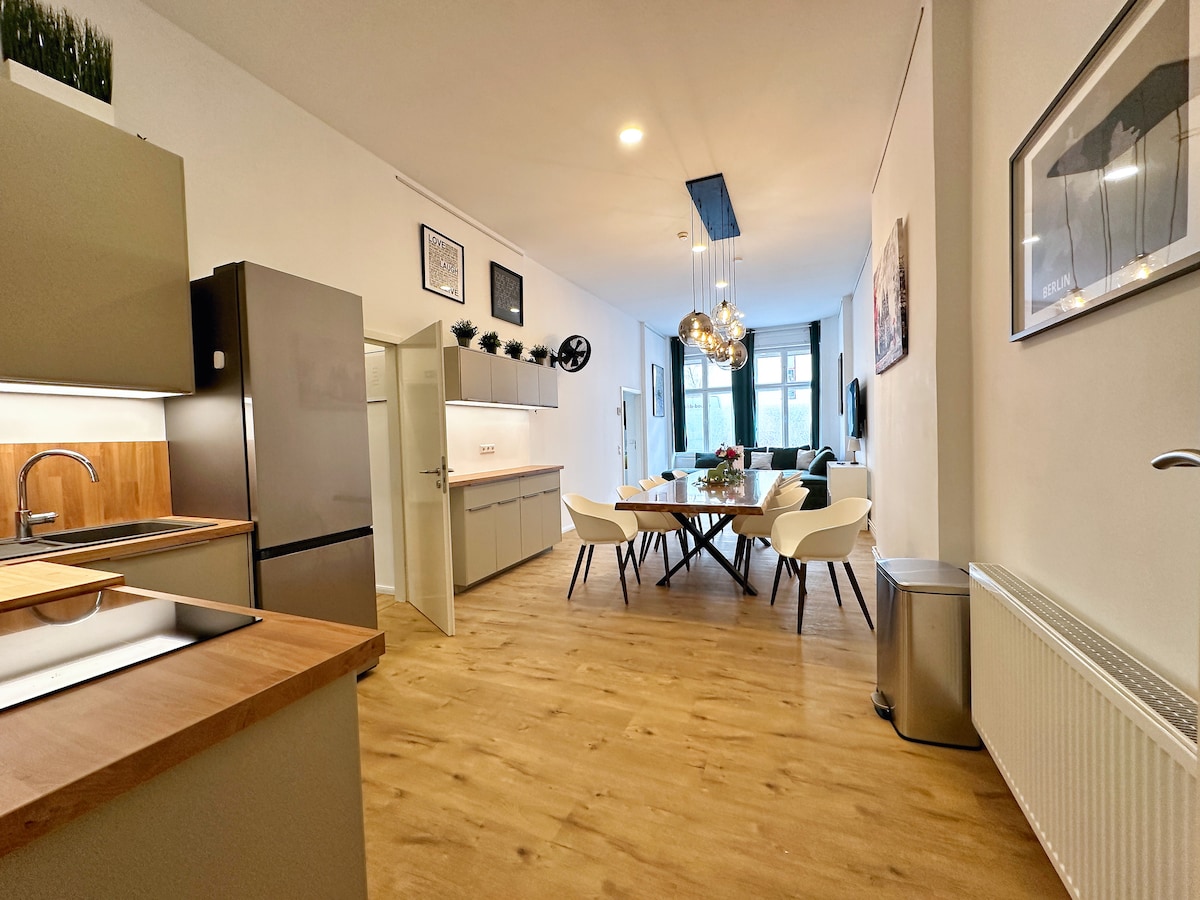 The kitchen features modern cabinetry and a stainless-steel refrigerator. A spacious dining table accommodates six, surrounded by white chairs. Natural light floods the area through large windows, enhancing the warm wood flooring and sophisticated atmosphere.