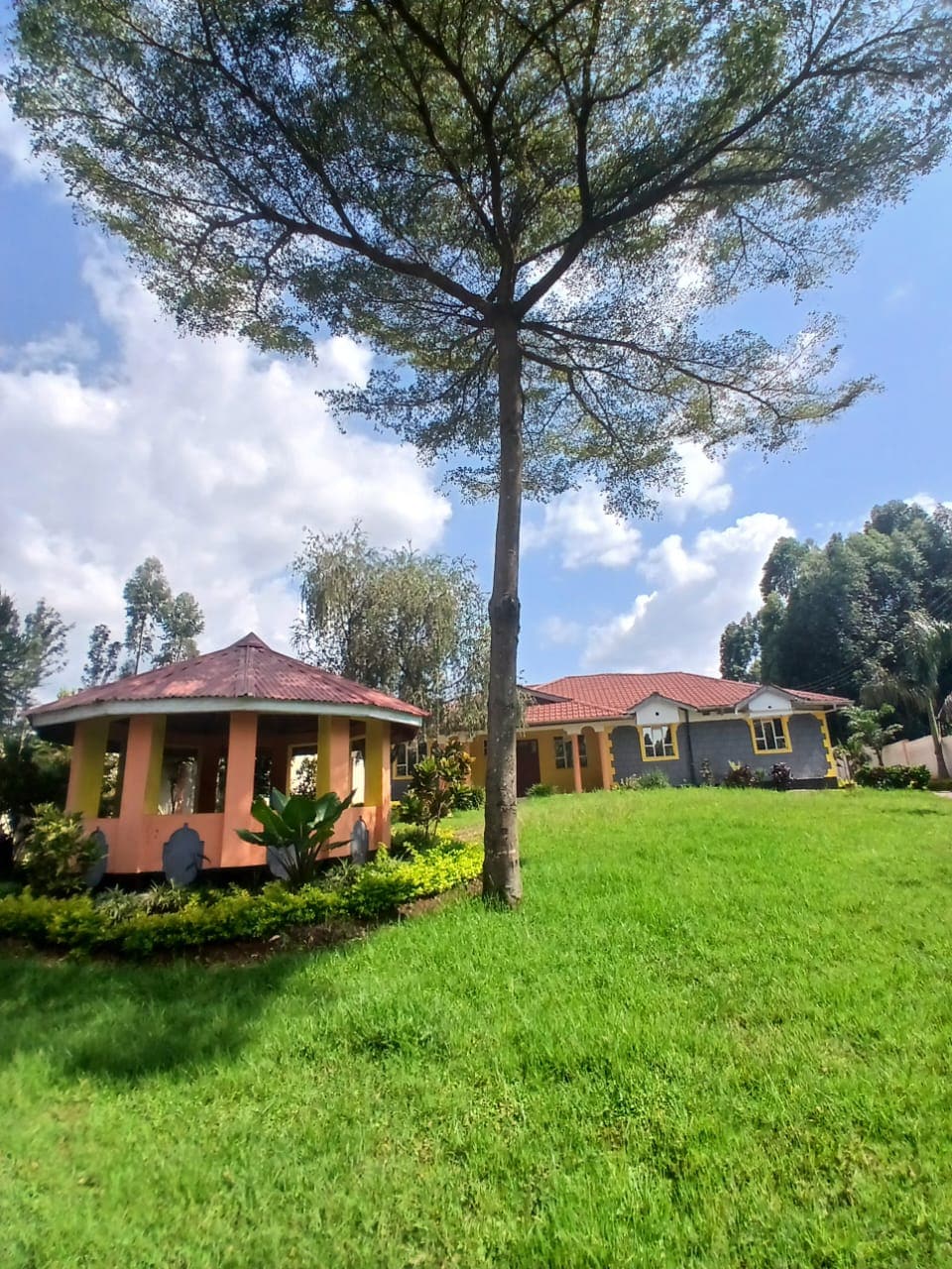 A charming property is situated on a lush green lawn, featuring a gazebo with a red roof and wooden columns. The main house, with a combination of stone and yellow walls, displays a welcoming entrance under the shade of a tall tree.