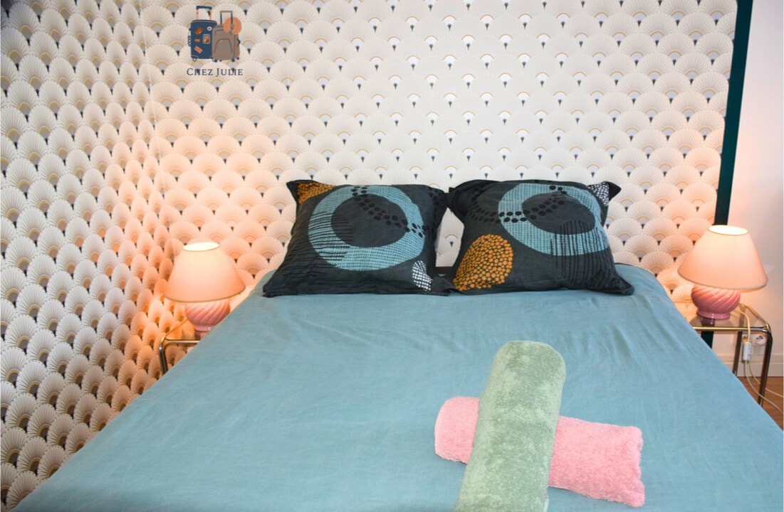 A sleeping area features a spacious bed with two decorative pillows placed against a textured headboard. Soft lighting is provided by two bedside lamps. A rolled towel lies on the bed, adding a touch of color to the serene environment.