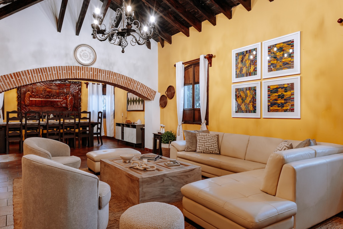 A spacious living area is highlighted by a neutral-colored sectional sofa and a wooden coffee table. Art pieces are displayed on the walls, complementing the warm yellow backdrop. Exposed wooden beams and a chandelier add character to the room, which features natural light from large windows.