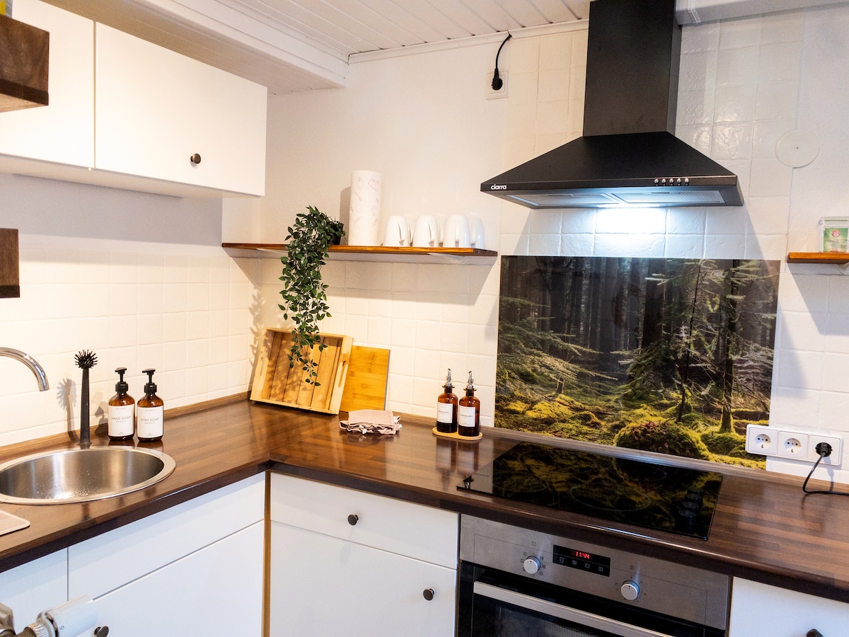 The kitchen features modern appliances and ample counter space. A stainless-steel sink is positioned near a sleek stove. Shelves display decorative items along with essential cooking products. A nature-themed backsplash adds a touch of tranquility to the functional area.