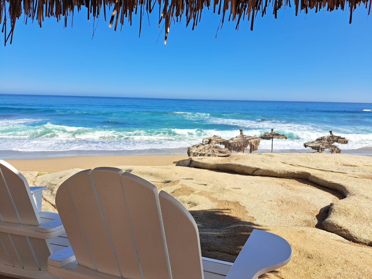A sandy beach scene is visible, with gentle waves lapping at the shore. Two white Adirondack chairs provide seating, positioned to face the ocean. Thatched umbrellas are set up in the background, adding to the coastal atmosphere under a clear blue sky.