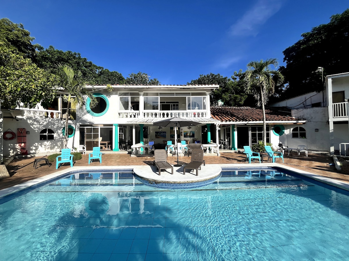 A tranquil outdoor area features a large swimming pool bordered by sun loungers. The two-story structure is visible in the background, with expansive glass windows reflecting natural light. Lush greenery surrounds the space, enhancing the serene environment.