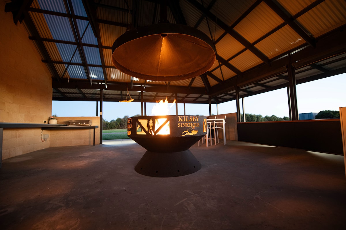 A central firepit with a large circular design is surrounded by a spacious, covered area. Bright flames emerge from the center, providing warmth and ambiance. The backdrop features large, open spaces with sliding doors that connect to the natural environment.