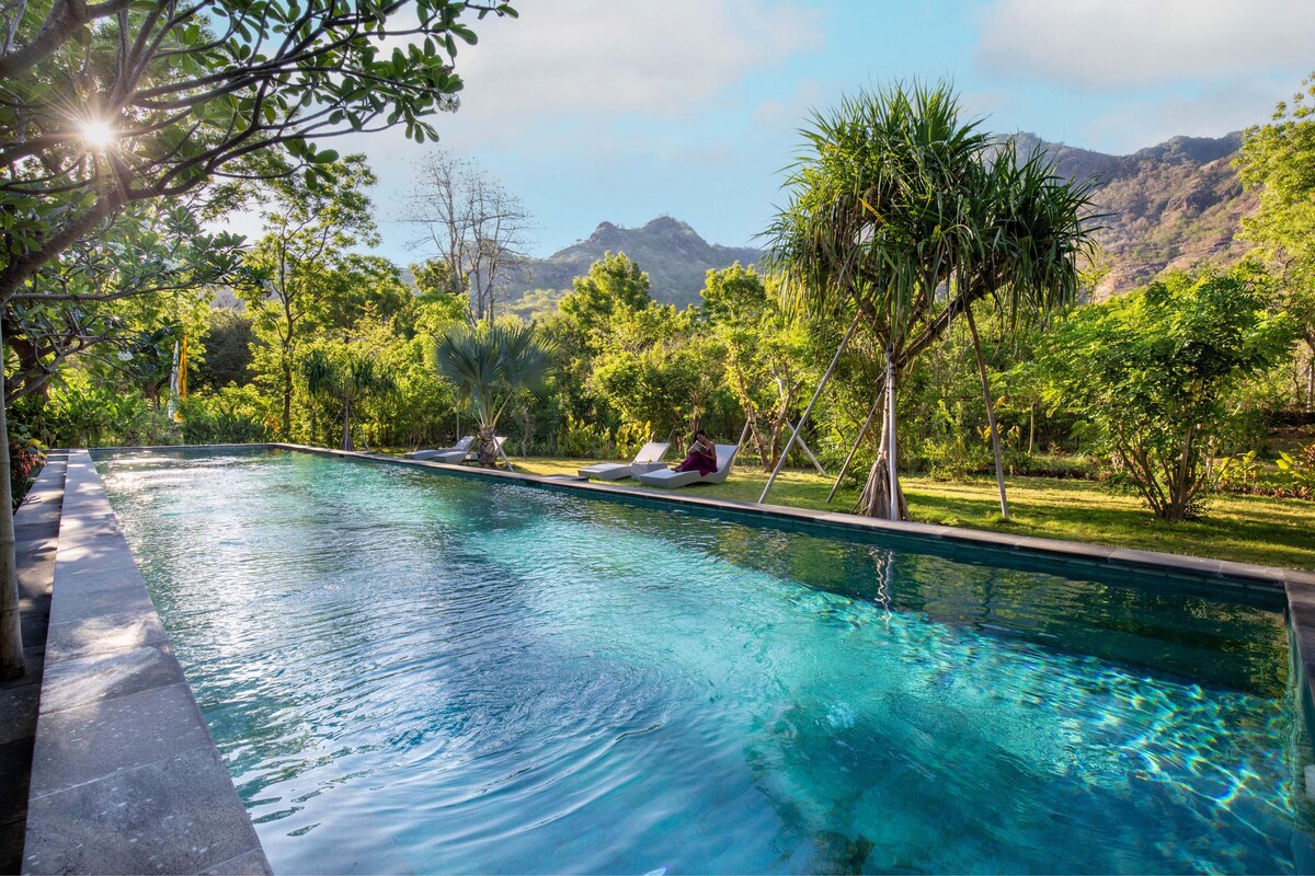 A serene swimming pool reflects the surrounding lush gardens and distant mountains. Sunlight filters through green foliage, creating a calm atmosphere. Lounge chairs are positioned beside the pool, inviting relaxation. The clear water of the pool enhances the tranquil setting.