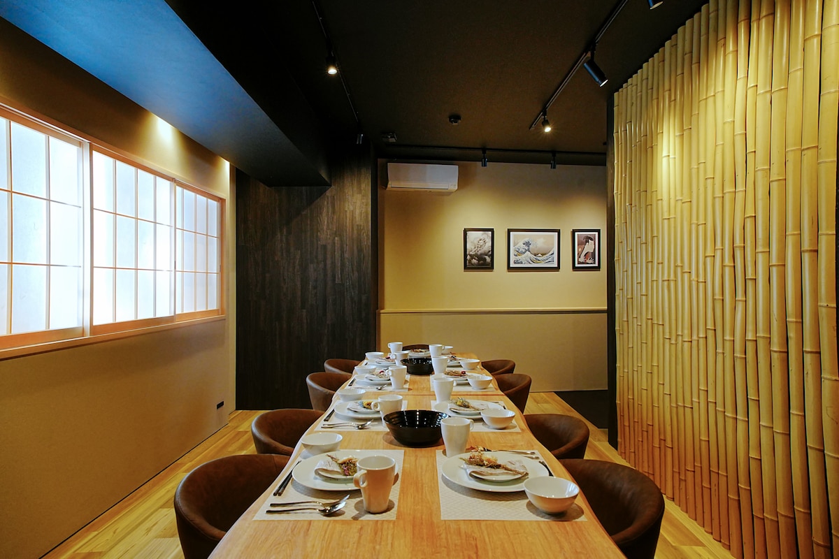 A dining area featuring a long wooden table set with dishware, surrounded by brown chairs. Soft lighting highlights bamboo accents along one wall, and framed artwork adds visual interest. Natural light filters through a window adorned with traditional shoji screens.