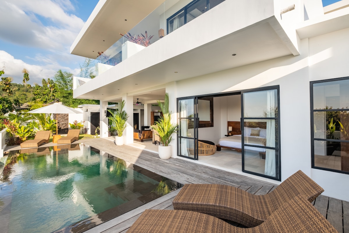 An elegant villa exterior features a private pool bordered by sun loungers and tropical greenery. Expansive glass doors open to the interior, allowing for a seamless connection between outdoor and indoor spaces while providing views of the serene surroundings.