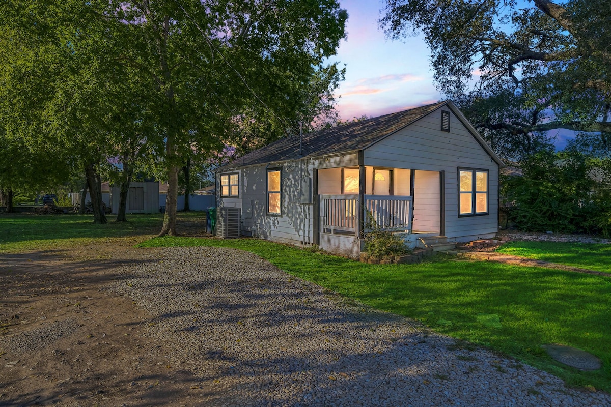 A cozy home exterior is shown at dusk, with warm light spilling from large windows. Surrounding trees provide shade in a well-maintained yard, and a gravel driveway leads to the entrance, creating an inviting outdoor space.