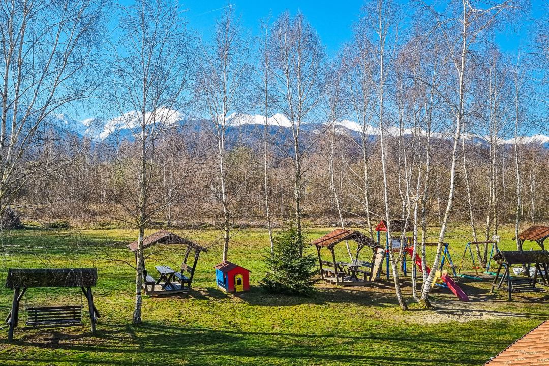 A spacious garden features several playground structures among tall trees. Picnic tables and swings are visible, with a small playhouse nearby. The background showcases mountains capped with snow under a clear blue sky, highlighting the serene natural setting.