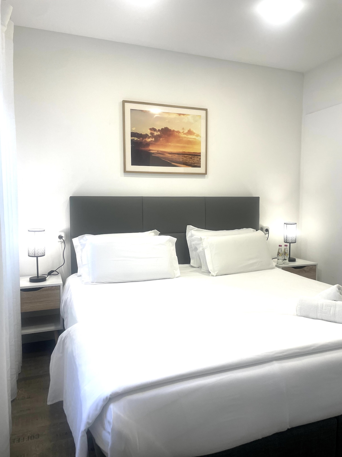 A bright bedroom features a spacious bed with crisp white linens and decorative pillows. Nightstands with bedside lamps are placed on either side, complementing a large artwork above the headboard. Soft natural light enters through a nearby window, enhancing the room's inviting ambiance.