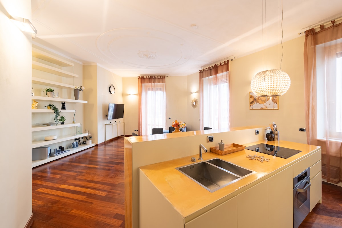 A spacious kitchen area features a modern island design with a sleek countertop. A double sink and modern appliances are visible. Natural light filters through two large windows, highlighting the warm wooden flooring and elegant shelving along the back wall.