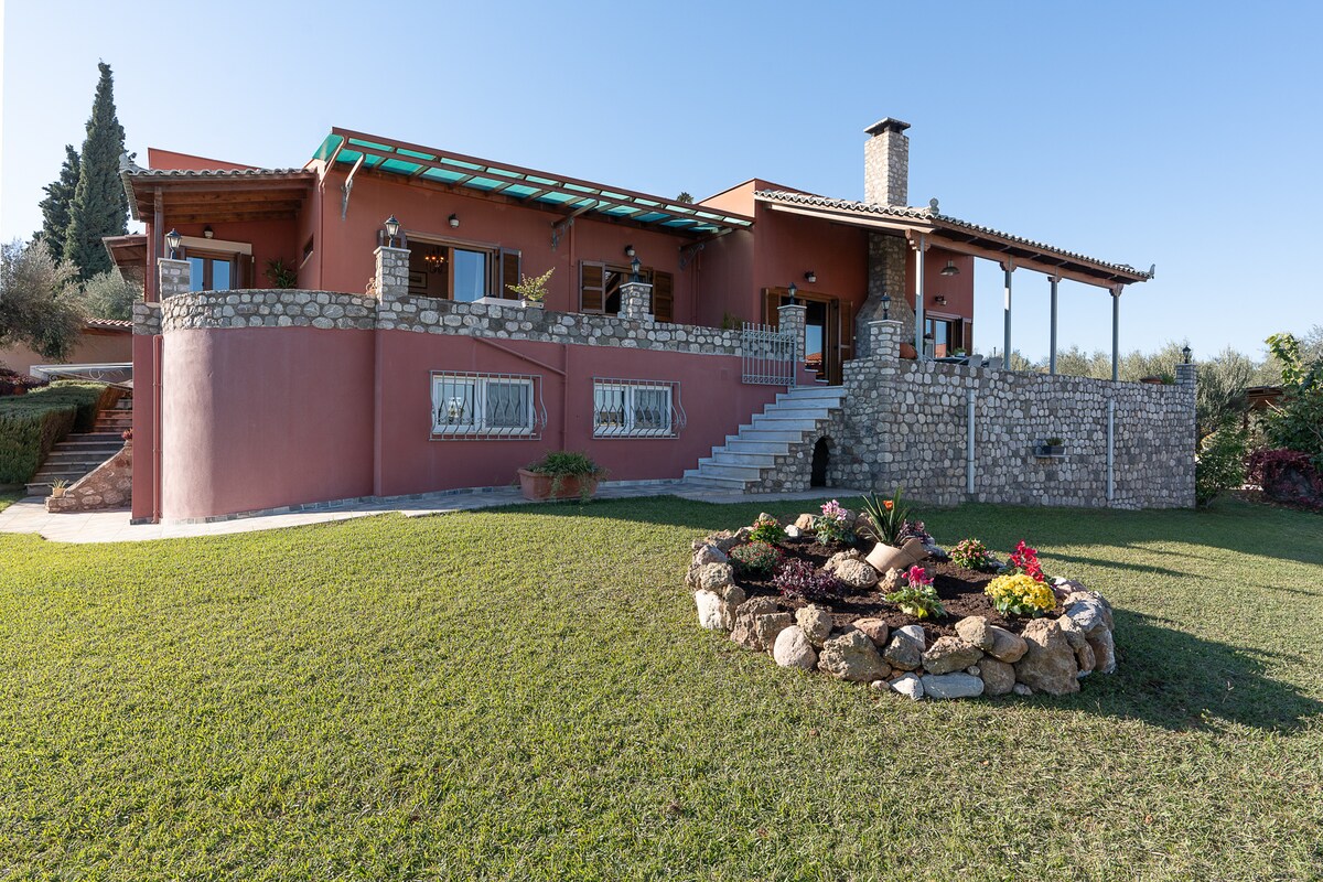 The exterior of the residence features a spacious layout with a combination of stone and smooth walls. A variety of plants and flowers surround a stone flowerbed at the forefront, and a cozy terrace with a chimney is visible, offering views of the lush surroundings.