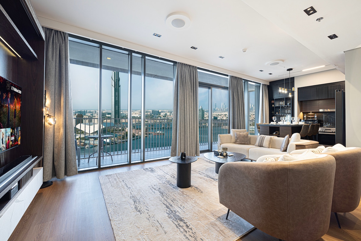 A stylish living area is presented with floor-to-ceiling windows, allowing natural light to fill the space. Comfortable seating is arranged around a chic coffee table, with a modern entertainment unit visible. An open kitchen area is seen in the background, providing a seamless flow.