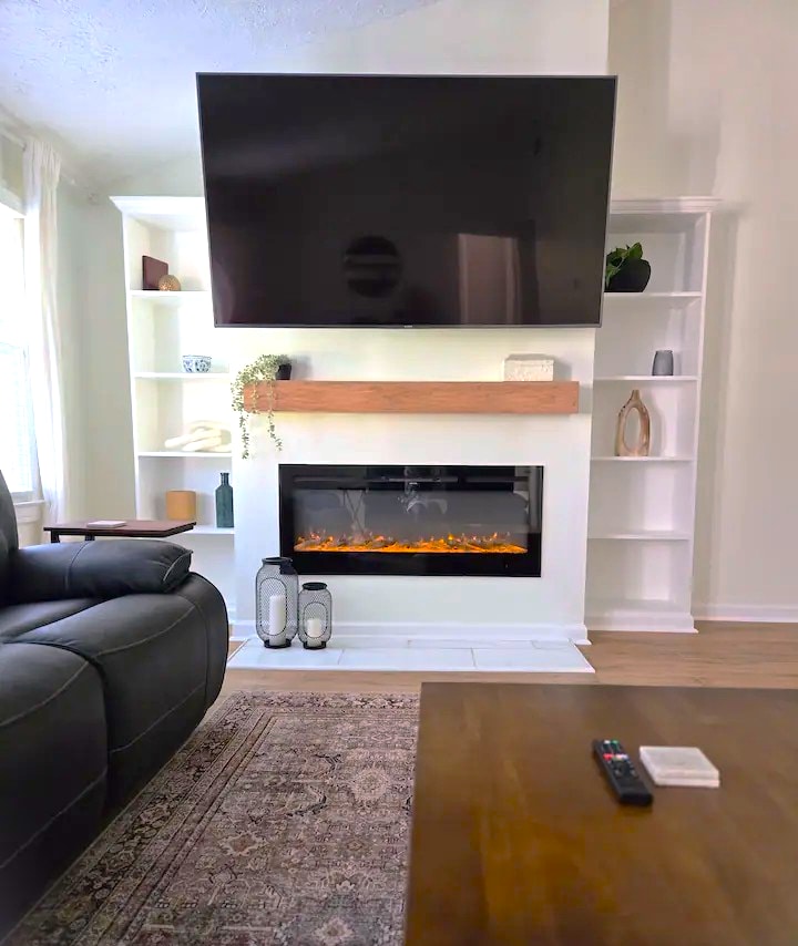 A modern living area features a wall-mounted television above a sleek electric fireplace. The stylish mantel is adorned with decorative items, while built-in shelves display a mix of curated objects. A cozy seating area includes a recliner, providing a comfortable spot to relax.