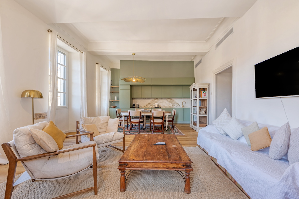 A spacious living area features natural light filtering through large windows. A wooden coffee table is surrounded by comfortable seating, including two white armchairs and a sofa. The open kitchen in soft green tones is visible in the background, creating a warm, inviting space.
