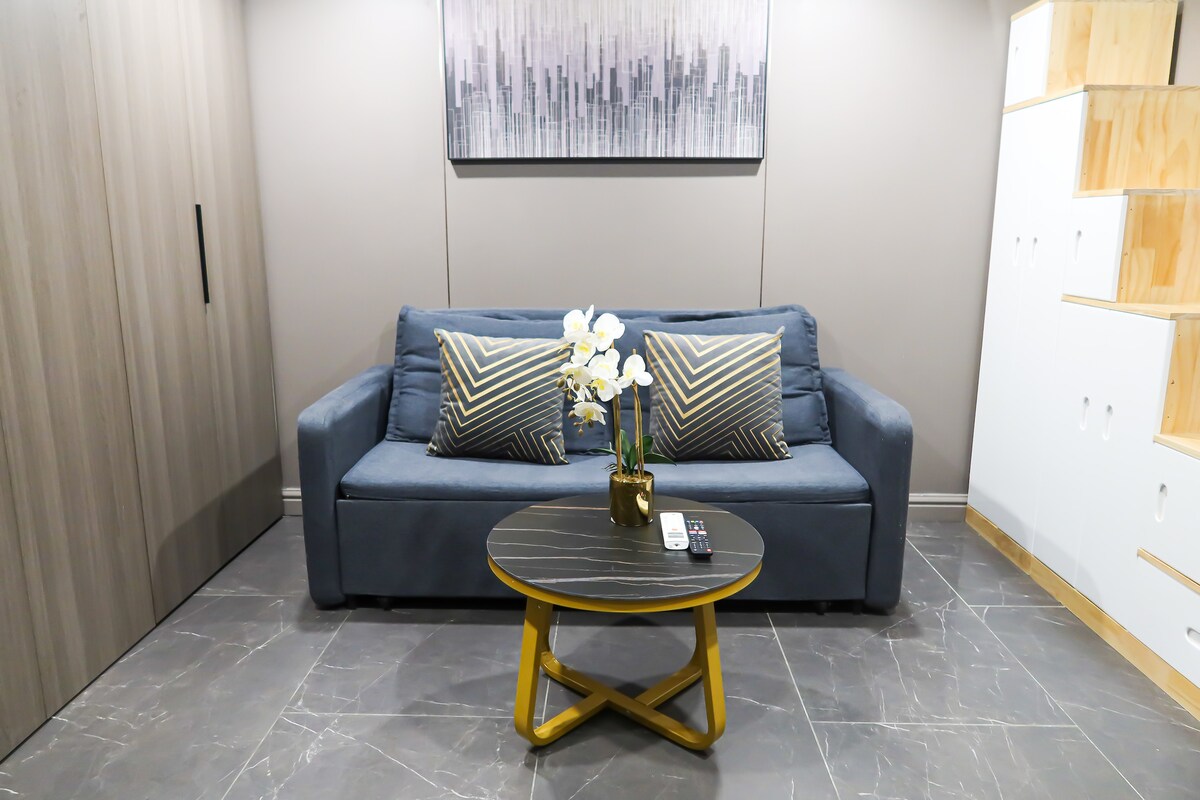 A cozy seating area features a two-seater sofa adorned with patterned cushions. A round coffee table sits in front, with a small vase of white flowers as a centerpiece. The walls are finished in neutral tones, and artwork is displayed above the sofa.