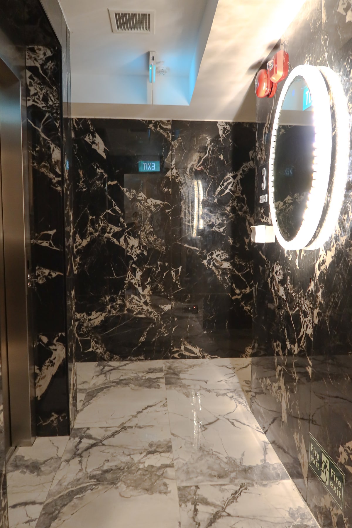 A modern corridor is showcased with dark marble walls and a polished marble floor. Illuminated by a circular mirror and ceiling lights, the space features a nearby elevator and an exit sign. The overall design adds a sleek and sophisticated touch.