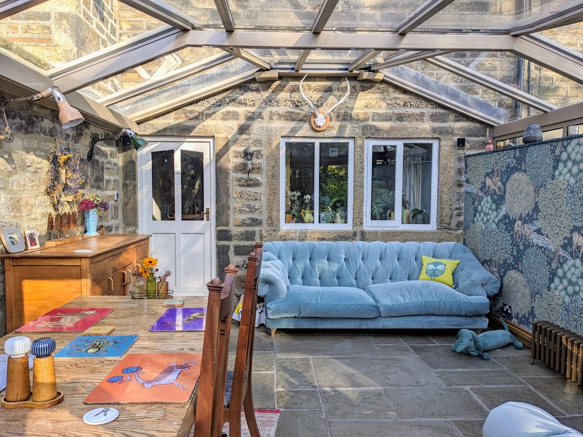 A conservatory features a large, soft blue sofa and a wooden dining table adorned with colorful placemats. Light streams through the glass roof, enhancing the stone walls and providing a bright and inviting space for relaxation and dining.