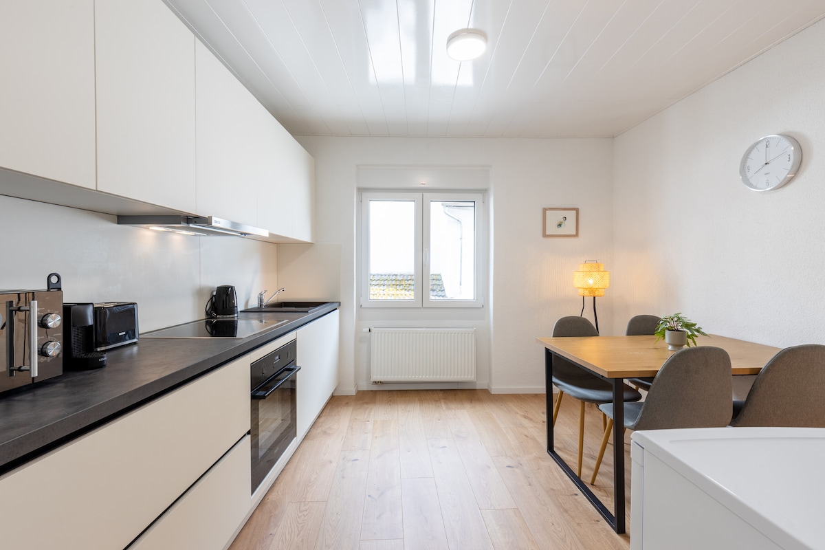 A modern kitchen features sleek white cabinetry and a dark countertop. Stainless steel appliances, including a microwave and coffee maker, are visible. A dining table with grey upholstered chairs is positioned nearby, illuminated by soft lighting. A window offers natural light, complemented by minimalist decor.